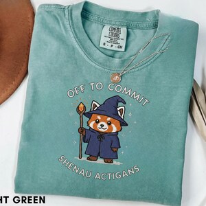 Może przedstawiać: Jasnozielona koszulka z napisem "OFF TO COMMIT SHENAU ACTIGANS" i ilustracją pandy rudej w czapce i szacie czarodzieja, trzymającej laskę. Srebrny naszyjnik z wisiorkiem leży na koszulce.