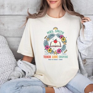 May include: A cream-colored t-shirt featuring a graphic with the text "MRS. SMITH", "TEACH LOVE INSPIRE", and "Back to School 2026". The design includes an open book with a red apple, surrounded by tropical leaves, hibiscus flowers, and starfish.