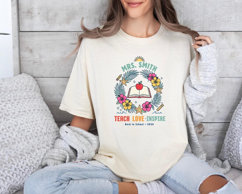 May include: A cream-colored t-shirt featuring a graphic with the text "MRS. SMITH", "TEACH LOVE INSPIRE", and "Back to School 2026". The design includes an open book with a red apple, surrounded by tropical leaves, hibiscus flowers, and starfish.