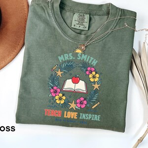 May include: A moss green crew neck t-shirt featuring a graphic of an open book with a red apple on top, surrounded by a wreath of palm leaves, hibiscus flowers, starfish, a pencil, and a ruler. The text reads MRS. SMITH, TEACH LOVE INSPIRE, BACK TO SCHOOL 2024.