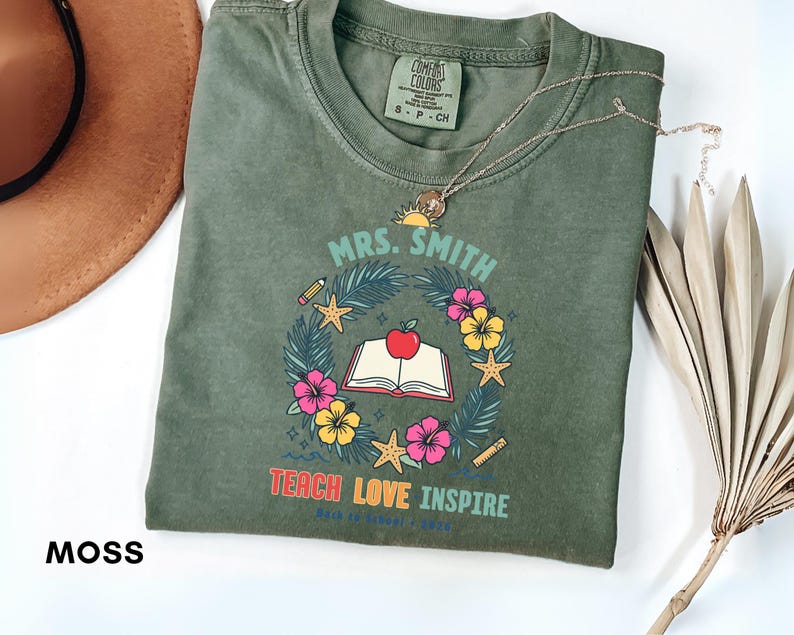 May include: A moss green crew neck t-shirt featuring a graphic of an open book with a red apple on top, surrounded by a wreath of palm leaves, hibiscus flowers, starfish, a pencil, and a ruler. The text reads MRS. SMITH, TEACH LOVE INSPIRE, BACK TO SCHOOL 2024.