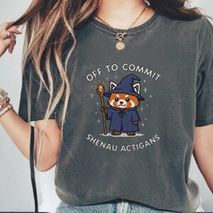 Może przedstawiać: Ciemnoszara koszulka z kresk&oacute;wkowym czerwonym pandą ubranym jako czarodziej, trzymającym laskę. Tekst "OFF TO COMMIT SHENAU ACTIGANS" jest wydrukowany nad i pod ilustracją. Koszulka ma kr&oacute;tkie rękawy i okrągły dekolt.