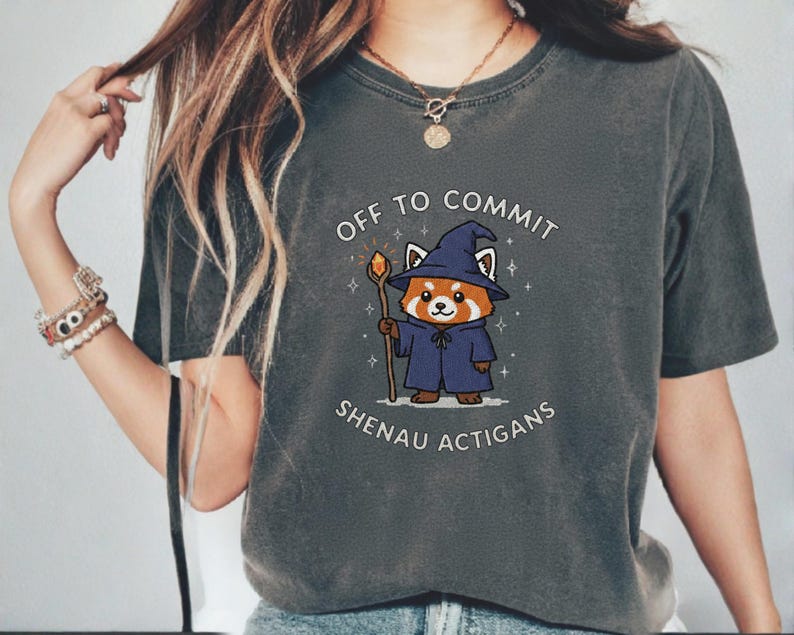 Może przedstawiać: Ciemnoszara koszulka z kresk&oacute;wkowym czerwonym pandą ubranym jako czarodziej, trzymającym laskę. Tekst "OFF TO COMMIT SHENAU ACTIGANS" jest wydrukowany nad i pod ilustracją. Koszulka ma kr&oacute;tkie rękawy i okrągły dekolt.