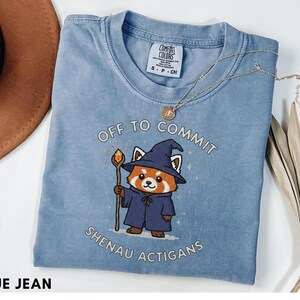 Może przedstawiać: Niebieska koszulka Comfort Colors z napisem "OFF TO COMMIT SHENAU ACTIGANS" i grafiką kresk&oacute;wkowego czerwonego pandy-czarodzieja. Czerwona panda ma na sobie niebieski kapelusz i szatę czarodzieja, trzymając laskę z pomarańczową kulą.