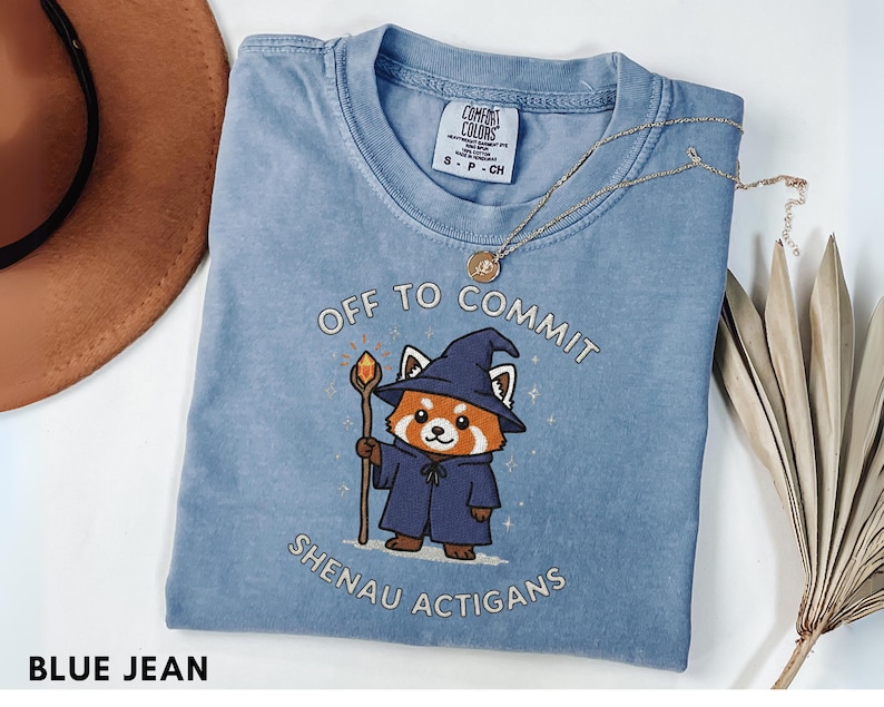 Może przedstawiać: Niebieska koszulka Comfort Colors z napisem "OFF TO COMMIT SHENAU ACTIGANS" i grafiką kresk&oacute;wkowego czerwonego pandy-czarodzieja. Czerwona panda ma na sobie niebieski kapelusz i szatę czarodzieja, trzymając laskę z pomarańczową kulą.