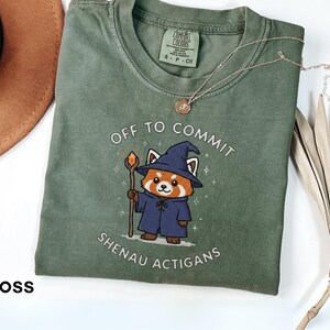 Może przedstawiać: Zielona koszulka w kolorze mchu z napisem "OFF TO COMMIT SHENAU ACTIGANS" i kresk&oacute;wkową czerwoną pandą w stroju czarodzieja. Widoczny jest r&oacute;wnież złoty naszyjnik i brązowy kapelusz.