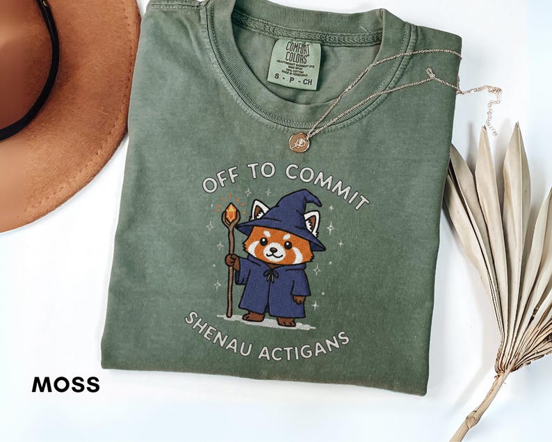 Może przedstawiać: Zielona koszulka w kolorze mchu z napisem "OFF TO COMMIT SHENAU ACTIGANS" i kresk&oacute;wkową czerwoną pandą w stroju czarodzieja. Widoczny jest r&oacute;wnież złoty naszyjnik i brązowy kapelusz.