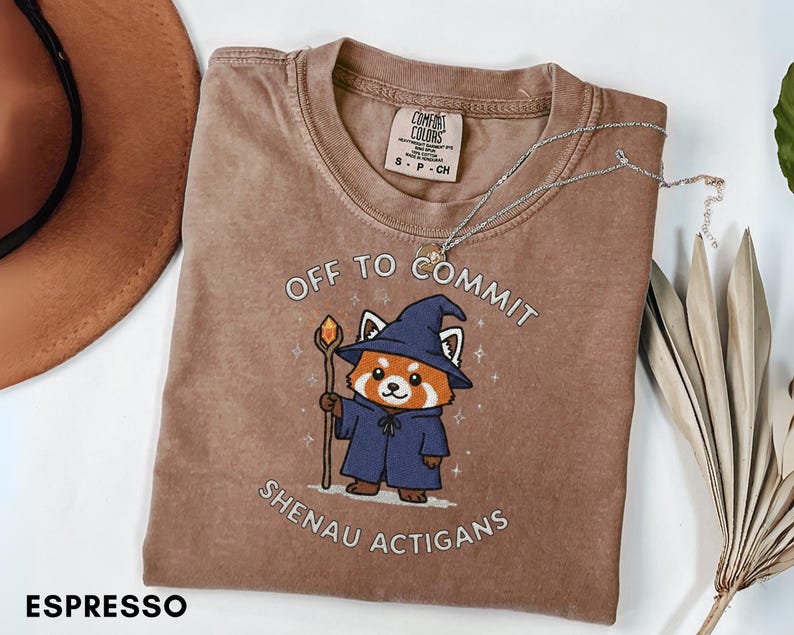 Może przedstawiać: Koszulka w kolorze espresso z grafiką czerwonej pandy-czarodzieja trzymającego laskę. Tekst na koszulce brzmi "OFF TO COMMIT SHENAU ACTIGANS". Srebrny naszyjnik jest zarzucony na koszulkę. Koszulka jest złożona na białej powierzchni.