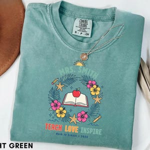 May include: A light green t-shirt featuring a graphic with the text "MRS. SMITH", "TEACH LOVE INSPIRE", and "Back to School 2026". The design includes a central open book with an apple, surrounded by a wreath of tropical leaves, flowers, and starfish.