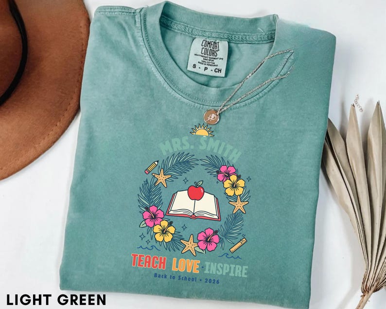 May include: A light green t-shirt featuring a graphic with the text "MRS. SMITH", "TEACH LOVE INSPIRE", and "Back to School 2026". The design includes a central open book with an apple, surrounded by a wreath of tropical leaves, flowers, and starfish.
