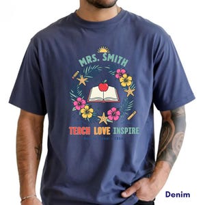 May include: A denim blue short-sleeve t-shirt featuring a graphic of an open book with a red apple, surrounded by a wreath of tropical flowers, starfish, and pencils. The text reads MRS. SMITH, TEACH LOVE INSPIRE, and 2026 in colorful lettering.