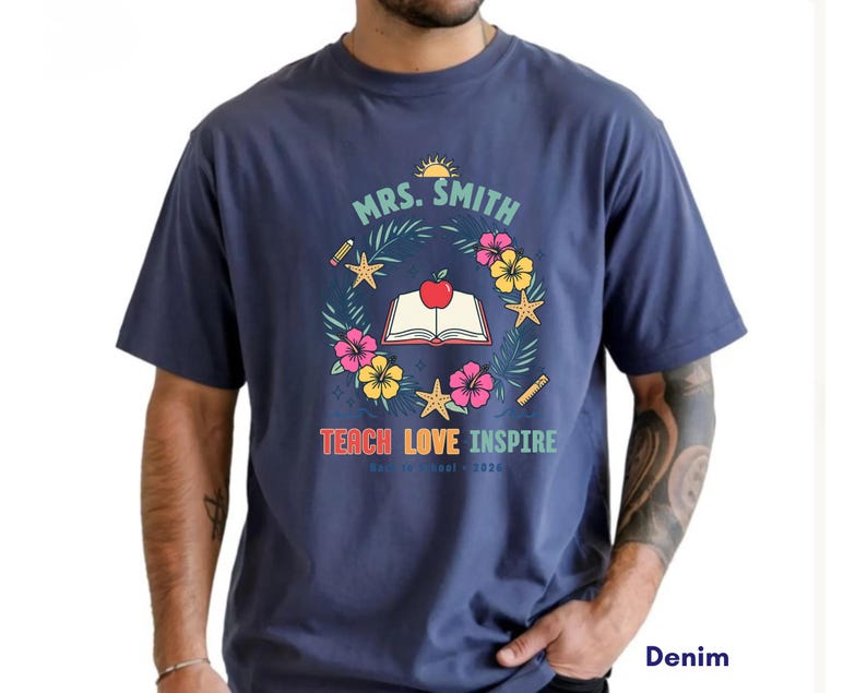 May include: A denim blue short-sleeve t-shirt featuring a graphic of an open book with a red apple, surrounded by a wreath of tropical flowers, starfish, and pencils. The text reads MRS. SMITH, TEACH LOVE INSPIRE, and 2026 in colorful lettering.