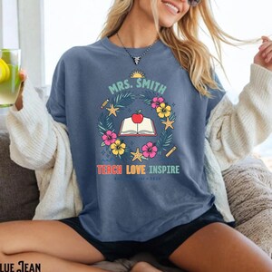 May include: A blue denim-colored t-shirt featuring a graphic print with the text "MRS. SMITH" and "TEACH LOVE INSPIRE". The design includes a central open book with a red apple, surrounded by a wreath of tropical flowers, leaves, and starfish.