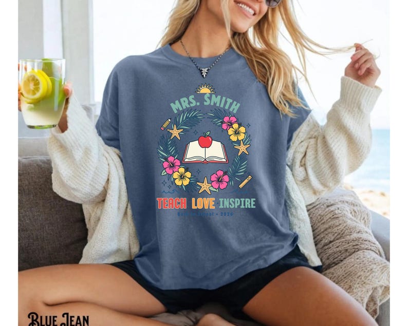 May include: A blue denim-colored t-shirt featuring a graphic print with the text "MRS. SMITH" and "TEACH LOVE INSPIRE". The design includes a central open book with a red apple, surrounded by a wreath of tropical flowers, leaves, and starfish.