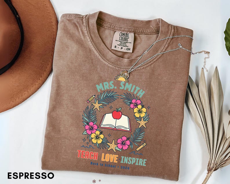 May include: An espresso-colored t-shirt featuring a graphic of an open book with a red apple on top, surrounded by a wreath of tropical leaves, hibiscus flowers, starfish, a pencil, and a ruler. The text reads MRS. SMITH, TEACH LOVE INSPIRE.