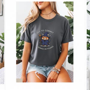 Może przedstawiać: Ciemnoszara koszulka z napisem "OFF TO COMMIT SHENAU ACTIGANS" i kresk&oacute;wkową czerwoną pandą w czapce i szacie czarodzieja. Panda trzyma laskę. Koszulka jest z kr&oacute;tkim rękawem.