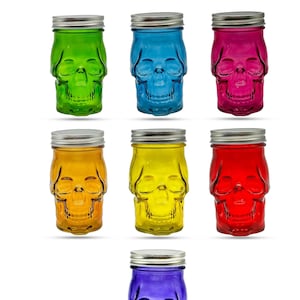 May include: Set of seven glass jars with skull designs. The jars are in various colors, including red, blue, green, yellow, orange, and purple. Each jar has a silver lid.