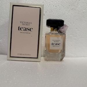 Könnte beinhalten: Ein Victoria's Secret Tease Eau de Parfum Flakon mit 100 ml Inhalt und die dazugehörige rechteckige Schachtel. Der Flakon besteht aus klarem Glas mit schwarzem Verschluss, goldenem Hals und hellrosa Flüssigkeit. Die Schachtel ist weiß.