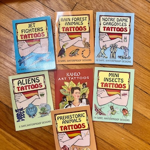 May include: A collection of eight colorful temporary tattoo books featuring various themes, including jet fighters, rain forest animals, gargoyles, aliens, Kahlo art, mini insects, and prehistoric animals. Each book includes a variety of designs and states the number of safe, waterproof designs included.