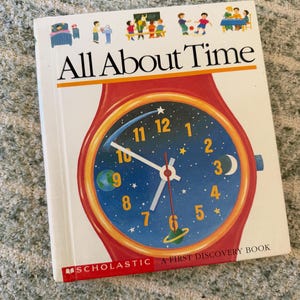 May include: A white hardcover book titled All About Time, A First Discovery Book by Scholastic. The cover features a large red wristwatch illustration with a blue space-themed clock face, yellow numbers, and small colorful illustrations of daily activities.
