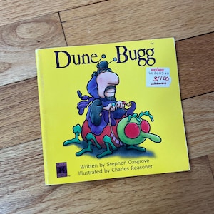 May include: A yellow children's book titled "Dune Bugg" with an illustration of a cartoon character riding a bug. The book cover features the text "Written by Stephen Cosgrove, Illustrated by Charles Reasoner."