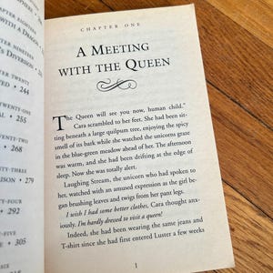 May include: Open book with text on a wooden surface. The page reads "A Meeting with the Queen" in large font, with the first paragraph beginning with a large drop cap. The text is from a fantasy novel.