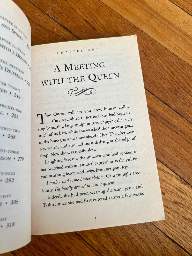 May include: Open book with text on a wooden surface. The page reads "A Meeting with the Queen" in large font, with the first paragraph beginning with a large drop cap. The text is from a fantasy novel.