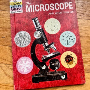 May include: A red book titled "The Microscope and What You See" from the "How and Why Wonder Book of" series. The cover features a black microscope with a variety of magnified images, including a fingerprint, a snowflake, a group of bacteria, and a beetle.