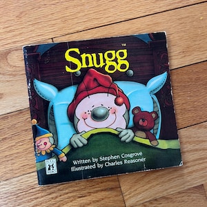 May include: A children's book titled "Snugg" with a cartoon illustration of a character in a red hat and a teddy bear. The book is written by Stephen Cosgrove and illustrated by Charles Reasoner. The cover is set against a wooden background.