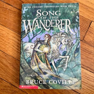 May include: A book cover for "Song of the Wanderer" by Bruce Coville, part of "The Unicorn Chronicles" series. The cover features a woman with a cat, a unicorn, and the text "Travel to the unknown". The cover is predominantly green and blue.