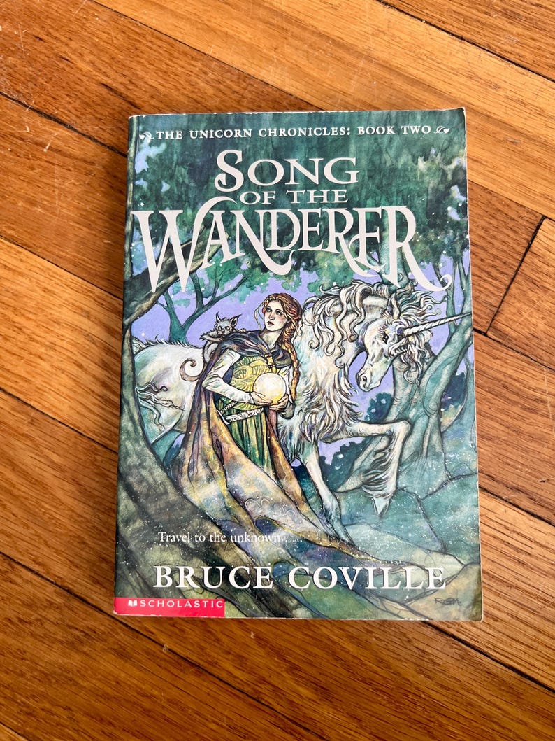 May include: A book cover for "Song of the Wanderer" by Bruce Coville, part of "The Unicorn Chronicles" series. The cover features a woman with a cat, a unicorn, and the text "Travel to the unknown". The cover is predominantly green and blue.