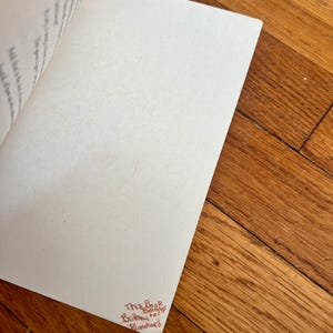May include: Open book with blank pages and text visible on the left page. The bottom right corner of the right page has handwritten text in red ink that reads "This Book Belongs to Brittany Blanchard". The book is on a wooden surface.