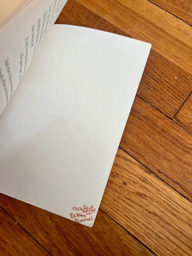 May include: Open book with blank pages and text visible on the left page. The bottom right corner of the right page has handwritten text in red ink that reads "This Book Belongs to Brittany Blanchard". The book is on a wooden surface.