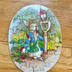May include: A wooden jigsaw puzzle featuring a cartoon rabbit wearing a blue jacket and holding a carrot. The rabbit is standing in a garden with a bird perched on a shovel. The puzzle is oval shaped and has a white background with green and brown accents. The puzzle is made by C.F.W. & Co.
