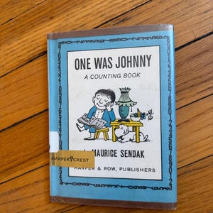 May include: A hardcover children's book titled One Was Johnny: A Counting Book by Maurice Sendak. The cover features a blue border with numbers, a central illustration of a child reading, a lamp, a bowl of fruit, and a dog, with Harper Crest text.