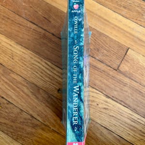 May include: A paperback book titled "Song of the Wanderer" by Coville. The book has a teal and blue cover with the word "Apple" and a pink apple logo at the top. The book is wrapped in clear plastic.