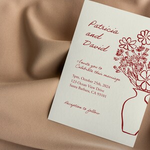 May include: Wedding invitation with a red floral illustration and the text "Patricia and David Invite you to Celebrate their marriage 5pm, October 25th, 2024 123 Ocean View Drive Santa Barbara, CA 93101 Reception to follow"