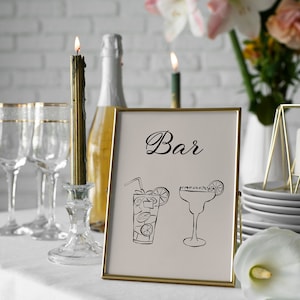 K&ouml;nnte beinhalten: Ein goldfarbener Rahmen mit dem Wort "Bar" in schwarzer Kursivschrift. Das Schild zeigt Illustrationen einer Margarita und eines Cocktails mit einem Strohhalm und einem Limettenkeil. Das Schild steht auf einem wei&szlig;en Tischtuch mit einer Kerze und Gl&auml;sern im Hintergrund.