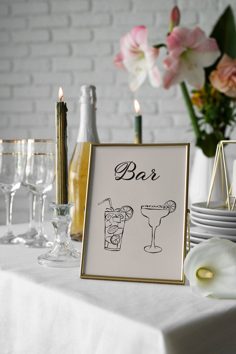 K&ouml;nnte beinhalten: Ein goldfarbener Rahmen mit dem Wort "Bar" in schwarzer Kursivschrift. Das Schild zeigt Illustrationen einer Margarita und eines Cocktails mit einem Strohhalm und einem Limettenkeil. Das Schild steht auf einem wei&szlig;en Tischtuch mit einer Kerze und Gl&auml;sern im Hintergrund.