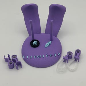 May include: A purple 3D printed rabbit head display stand with two long ears, a black button eye, a light blue stitched eye, and a light blue stitched mouth. The set includes two small purple clips and two purple clips with clear plastic loops.