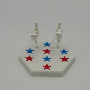 May include: A white hexagonal display stand featuring two vertical support posts with adjustable clear loops. The base is decorated with a pattern of small, flat, five-pointed stars in alternating red and blue colours across the top surface.