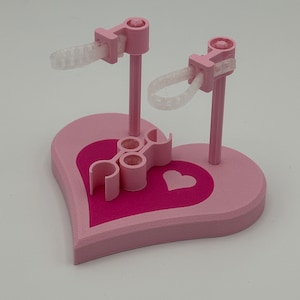 May include: A pink heart-shaped display stand featuring a darker pink heart inlay. The base includes two vertical posts with adjustable clear plastic loops at the top and a central pink clip component with two circular openings for holding small items.