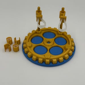 May include: A circular gold gear-shaped base with a blue centre featuring five blue circular inserts. Two vertical gold posts extend upwards, each holding a translucent white loop. Two separate gold C-shaped clips are positioned to the left.