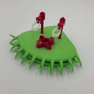 May include: A 3D printed green, semi-circular object with pointed, thorn-like protrusions along its curved edge. It features two upright magenta posts with attached white loops and a central magenta connector piece with four circular openings.