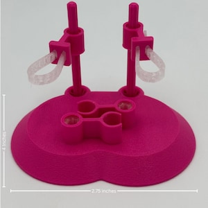 May include: A bright pink doll stand with a heart-shaped base, two vertical support poles, and two clear elastic loops. The stand features a central clip mechanism and measures 4 inches in height and 2.75 inches in width as indicated.