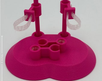 May include: A bright pink doll stand with a heart-shaped base, two vertical support poles, and two clear elastic loops. The stand features a central clip mechanism and measures 4 inches in height and 2.75 inches in width as indicated.