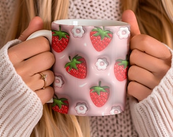 May include: A white ceramic mug with a pink background featuring a pattern of red strawberries and white flowers. The mug has a white handle and is held by a person wearing a cream-colored sweater.