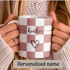 Puede incluir: Una taza de cerámica a cuadros rosa y blancos con el nombre "heather" escrito en cursiva y un pequeño diseño de corazón. Nombre personalizado.