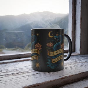 May include: Black ceramic mug featuring a blue serpent design with a crown, lotus flowers, and the text "THE INFINITE WISDOM". The mug has a black handle and a white interior. The background shows a window with a mountain view.