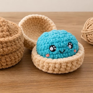 May include: A tan crocheted container with a lid and a matching open base holding a bright blue crocheted sphere with a smiling face, black eyes, and pink cheeks. The text La P'tite maille is visible in the bottom left corner of the image.
